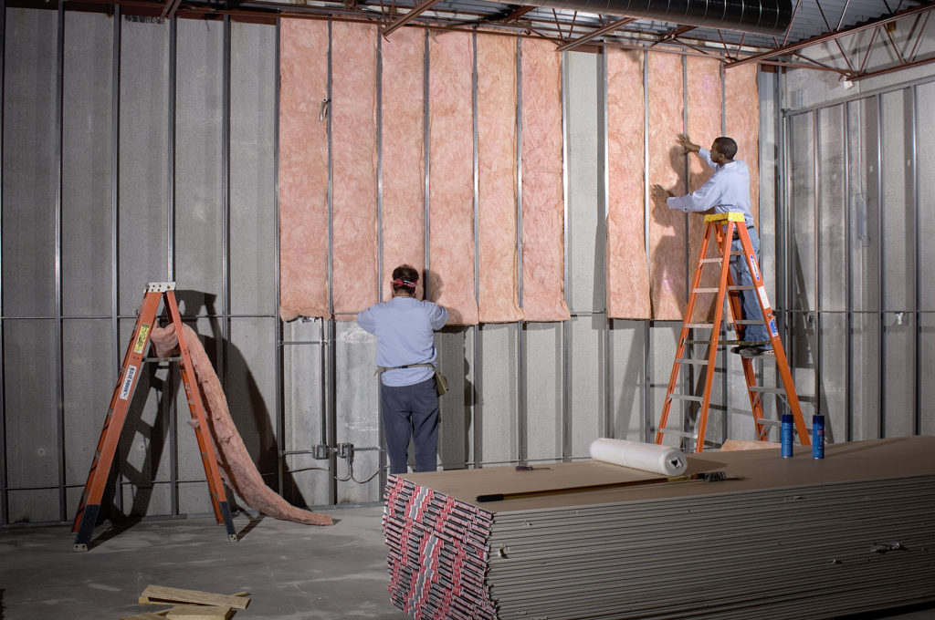 Two technicians installing pink fiberglass batt insulation in the wall of a large commercial building