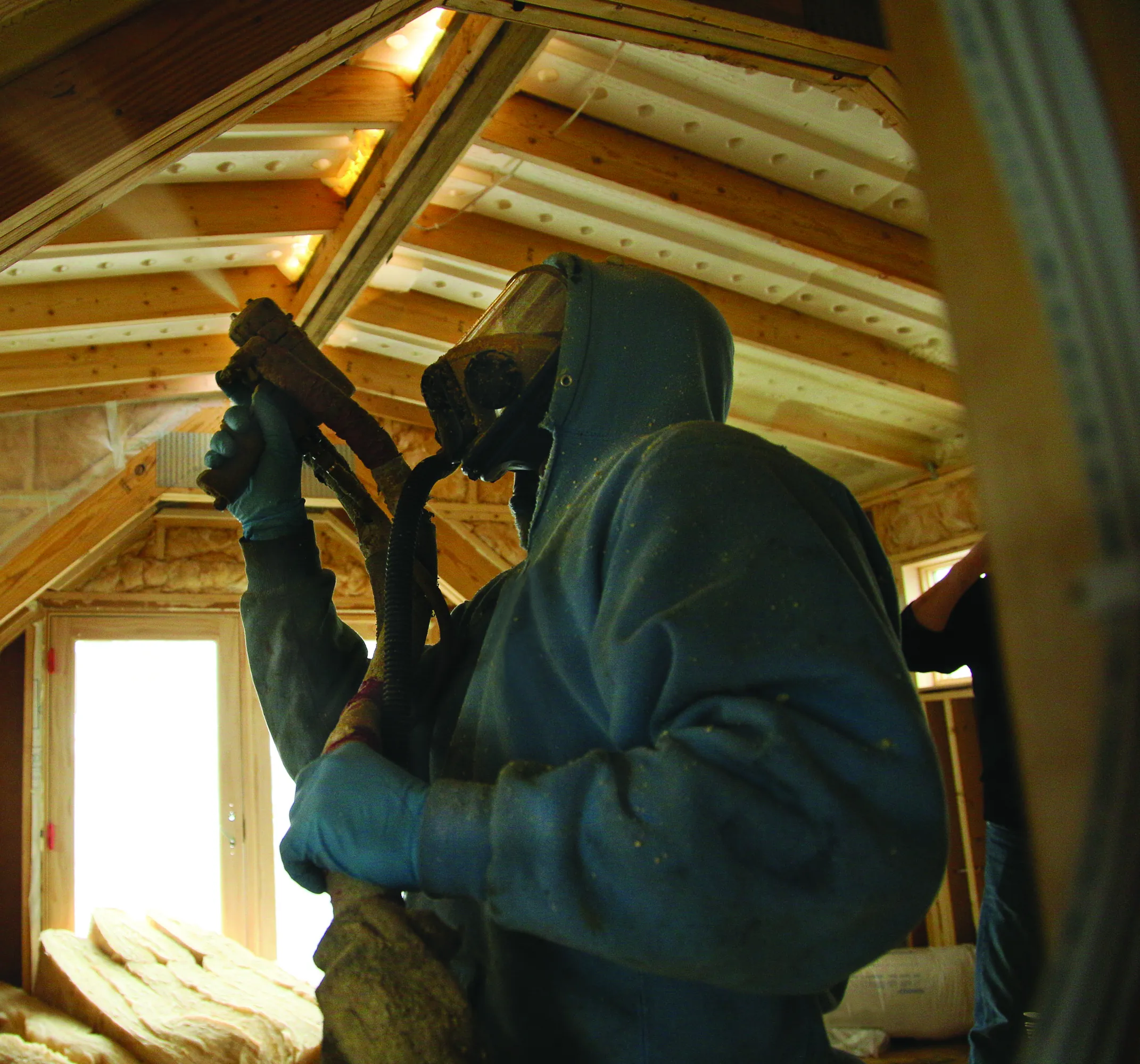spray foam insulation being installed by technician in hazmat suit, in ceiling of unfinished attic.